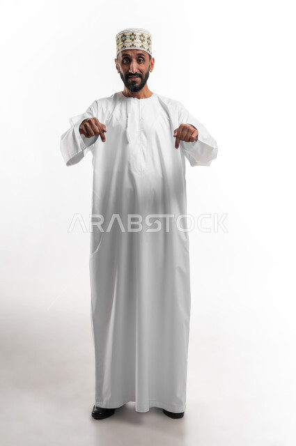 في هذه اللحظة (الآن), إيماءات تشير الى موقع معين, بورتريه  لرجل كبير السن عربي خليجي عماني يرتدي الدشداشة والكمه يشير بإصبعه نحو الأسفل وينظر إلى الكاميرا, حركات تدل على هذا المكان (هنا), صورة الجسم بالطول الكامل, خلفية بيضاء.