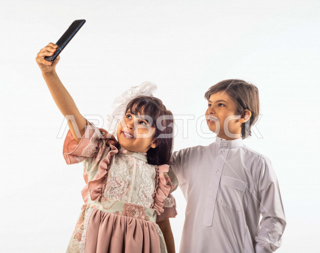 خلفية بيضاء لصبي وفتاه صغيران سعوديان مبتسمان ، تمسك الفتاه الجوال بيدها , يقومان بإلتقاط صور سيلفي تذكاريه بإستخدام الهاتف المحمول بإيماءات وجه ويدين مختلفة , بورتريه لطفل وطفلة سعوديان خليجيان سعيدان يرتديان لباس العيد ,الاحتفال بعيد الفطر السعيد