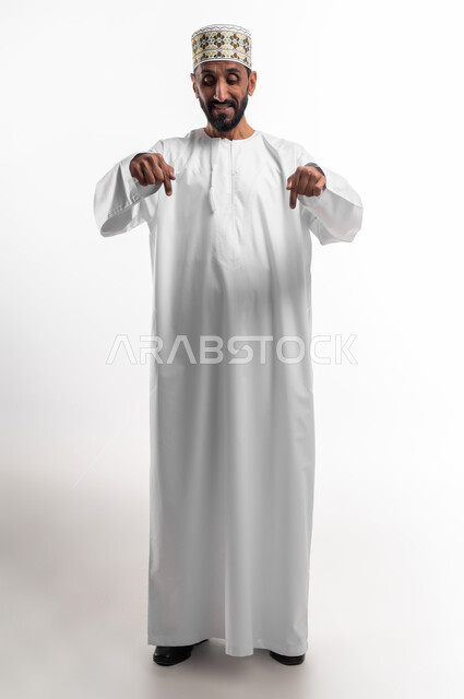 في هذه اللحظة (الآن), إيماءات تشير الى موقع معين, بورتريه  لرجل كبير السن عربي خليجي عماني يرتدي الدشداشة والكمه يشير بإصبعه نحو الأسفل وينظر إلى الأسفل, حركات تدل على هذا المكان (هنا), صورة الجسم بالطول الكامل, خلفية بيضاء.