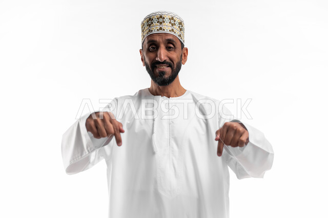 في هذه اللحظة (الآن), إيماءات تشير الى موقع معين, بورتريه مقرب لرجل كبير السن عربي خليجي عماني يرتدي الدشداشة والكمه يشير بإصبعه نحو الأسفل وينظر إلى الكاميرا, حركات تدل على هذا المكان (هنا), خلفية بيضاء.