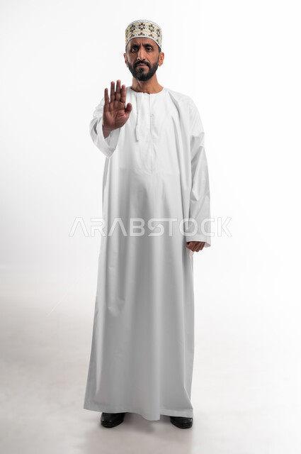 مفهوم الرفض والامتناع والانزعاج, بورتريه  لرجل كبير السن عربي خليجي عماني يرتدي الدشداشة والكمه  يمد يده نحو الأمام مع رفع الكف بإيماءات النهي عن فعل امر ما, طلب التوقف وعدم الاقتراب, تعابير الحزم والجدية, صورة الجسم بالطول الكامل,  خلفية بيضاء.