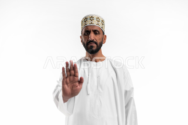 مفهوم الرفض والامتناع والانزعاج, بورتريه مقرب لرجل كبير السن عربي خليجي عماني يرتدي الدشداشة والكمه  يمد يده نحو الأمام مع رفع الكف بإيماءات النهي عن فعل امر ما, طلب التوقف وعدم الاقتراب, تعابير الحزم والجدية, خلفية بيضاء.
