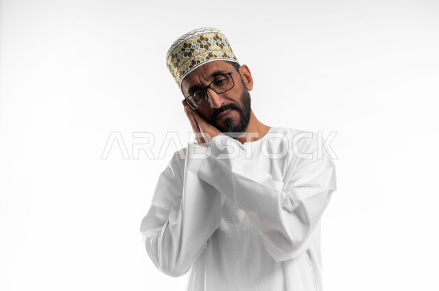 تعابير الشعور  بالنعاس والتعب, بورتريه مقرب لرجل عربي خليجي عماني يرتدي الدشداشة والكمه والنظارات الطبية يضع يديه تحت رأسه مع إغماض العينين  بإيماءات الحاجة الى النوم والراحة, خلفية بيضاء.