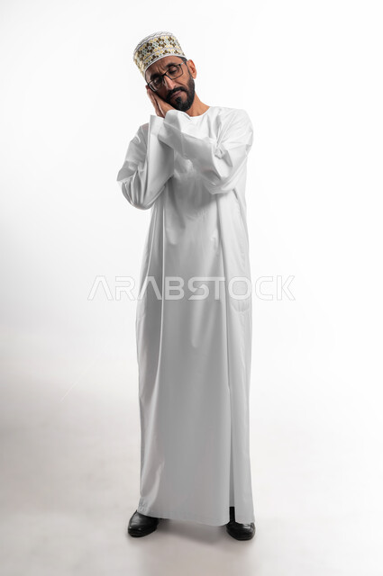 تعابير الشعور  بالنعاس والتعب, بورتريه لرجل عربي خليجي عماني يرتدي الدشداشة والكمه والنظارات الطبية يضع يديه تحت رأسه مع إغماض العينين  بإيماءات الحاجة الى النوم والراحة, صورة الجسم بالطول الكامل, خلفية بيضاء.