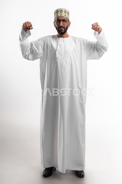 محاولة ضبط الأعصاب وعدم التوتر, بورتريه لرجل كبير السن عربي خليجي عماني يرتدي الدشداشة والكمه والنظارات الطبية يرفع قبضات يديه عاليا بإيماءات العصبية والانفعال, تعابير الغضب والانزعاج, صورة الجسم بالطول الكامل, خلفية بيضاء.