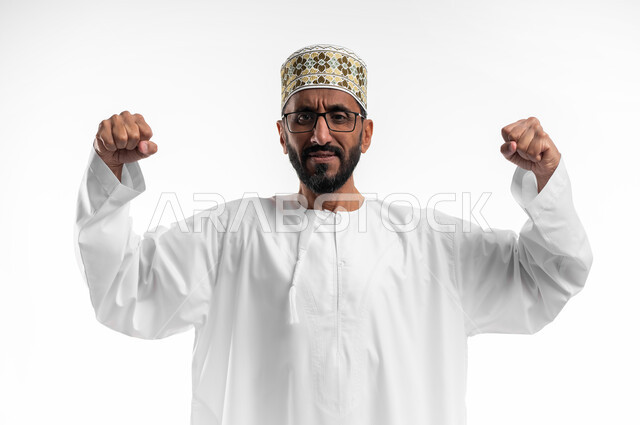 محاولة ضبط الأعصاب وعدم التوتر, بورتريه مقرب لرجل كبير السن عربي خليجي عماني يرتدي الدشداشة والكمه والنظارات الطبية  يرفع قبضات يديه عاليا بإيماءات العصبية والانفعال, تعابير الغضب والانزعاج, خلفية بيضاء.