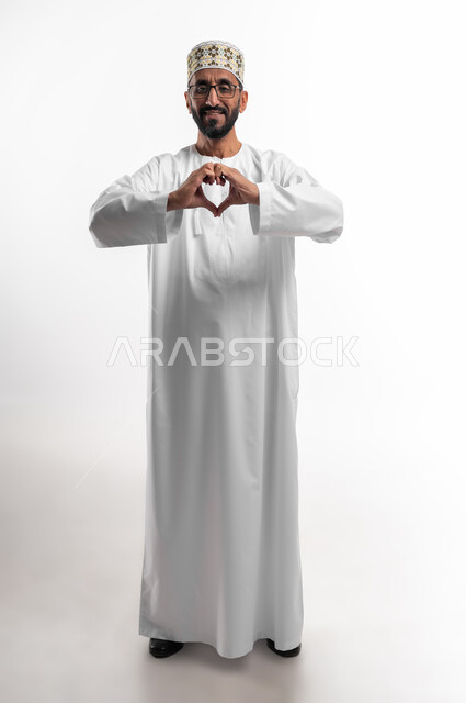 النظر للكاميرا بإيماءات الإعجاب والسرور، تشكيل قلب حب باليد، بورتريه لرجل عماني شايب يرتدي الدشداشة والكمه ونظارات طبية، تعابير فرح وابتسامة، الطول الكامل للجسم، خلفية بيضاء