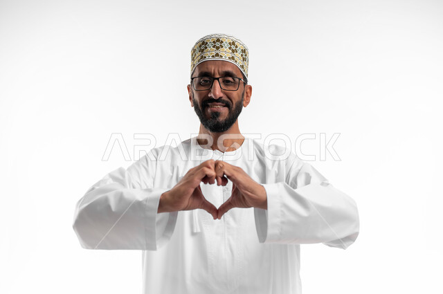 النظر للكاميرا بإيماءات الإعجاب والسرور، تشكيل قلب حب باليد، بورتريه لرجل عماني شايب يرتدي الدشداشة والكمه ونظارات طبية، تعابير فرح وابتسامة، الزي التقليدي للرجال في سلطنة عمان، خلفية بيضاء