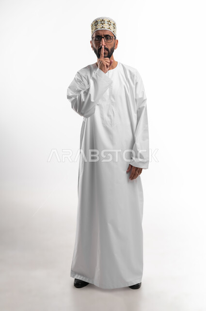 عقد الحاجبين و وضع إصبع السبابة على الفم إشارة لإنهاء الكلام، تعابير وجه تدل على الانزعاج، ايماءات تحذير وجدية، بورتريه لرجل عماني شايب يرتدي الدشداشة والكمه ونظارات طبية، التعصيب واعطاء التحذيرات، خلفية بيضاء