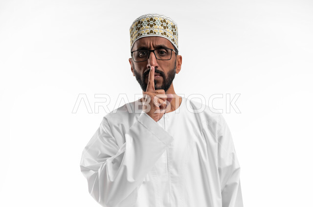 عقد الحاجبين و وضع إصبع السبابة على الفم إشارة لإنهاء الكلام، تعابير وجه تدل على الانزعاج، ايماءات تحذير وجدية، بورتريه لرجل عماني شايب يرتدي الدشداشة والكمه ونظارات طبية، التعصيب واعطاء التحذيرات، خلفية بيضاء