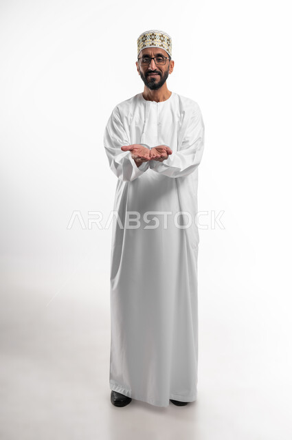 A hand movement indicating a request for help and assistance, extending the palms forward to indicate need, a portrait of an old Omani man wearing a dishdasha, a cuff, and medical glasses, the traditional dress for men in the Sultanate of Oman, gestures of helplessness and hope, white background.