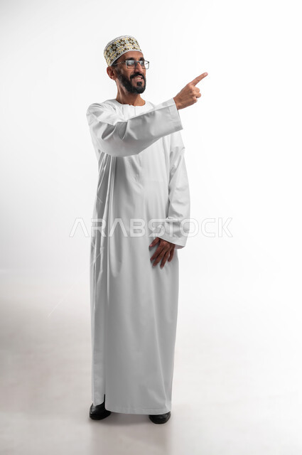 مد اليد إشارةً لشيءٍ ما بواسطة إصبع السبابة، شد الإنتباه ولفت النظر، ايماءات سعادة وفرح، بورتريه لرجل عماني شايب يرتدي الدشداشة والكمه ونظارات طبيه، الجسم بالطول الكامل، الزي التقليدي للرجال في سلطنة عمان، خلفيه بيضاء