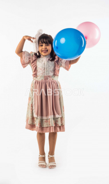 خلفية بيضاء لفتاه صغيرة سعودية خليجية مبتسمة وجميلة ، تنظر الى الكاميرا وتحمل بيدها  بالونين بلون أزرق وزهري بإيماءات وجه ويدين مختلفة تدل على السعادة , بورتريه لطفلة سعودية خليجية سعيدة ترتدي لباس العيد الانيق , الاحتفال بعيد الفطر السعيد ، عيد مبارك