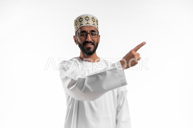 مد اليد إشارةً لشيءٍ ما بواسطة إصبع السبابة، شد الإنتباه ولفت النظر، ايماءات سعادة وفرح، بورتريه لرجل عماني شايب يرتدي الدشداشة والكمه ونظارات طبيه، الزي التقليدي للرجال في سلطنة عمان، خلفيه بيضاء