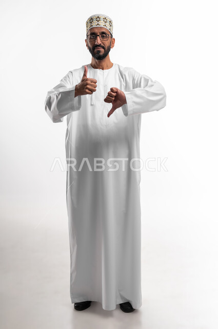 الموافقة وعدم الموافقة، بورتريه لرجل عماني شايب يرتدي الدشداشة والكمه ونظارات طبية، يرفع اصابع الابهام للاعلى وللاسفل بتعابير التردد في شيء ما, عدم الإعجاب و الرفض و القبول و الرضا, النظر الى الكاميرا بتعابير الحيرة، خلفية بيضاء