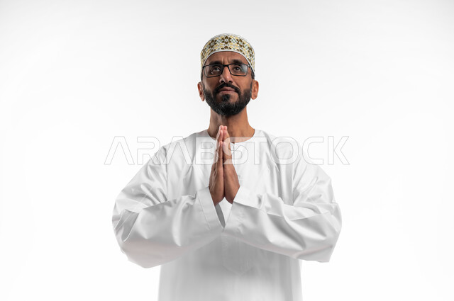 ضم الكفين امام الصدر للقيام بالدعاء، ايماءات التوسل والاسترحام، طلب المساعدة من المولى عز وجل، بورتريه لرجل عماني شايب يرتدي الدشداشة والكمه يدعي ويطلب المغفرة، خلفية بيضاء