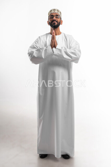 ضم الكفين امام الصدر للقيام بالدعاء، ايماءات التوسل والاسترحام، طلب المساعدة من المولى عز وجل، بورتريه لرجل عماني شايب يرتدي الدشداشة والكمه يدعي ويطلب المغفرة، خلفية بيضاء