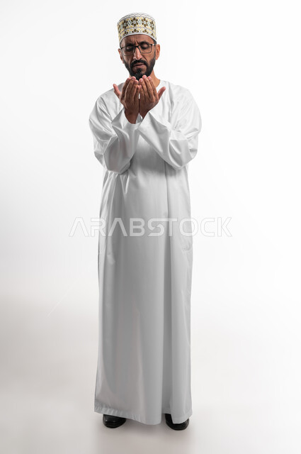 رفع اليدين للأعلى بهدف الدعاء والتضرع إلى الله تعالى،ايماءات التوسل والاسترحام، طلب المساعدة من المولى عز وجل، بورتريه لرجل عماني شايب يرتدي الدشداشة والكمه يدعي ويطلب المغفرة، خلفية بيضاء