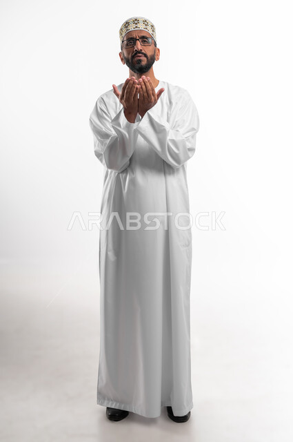 رفع اليدين للأعلى بهدف الدعاء والتضرع إلى الله تعالى،ايماءات التوسل والاسترحام، طلب المساعدة من المولى عز وجل، بورتريه لرجل عماني شايب يرتدي الدشداشة والكمه يدعي ويطلب المغفرة، خلفية بيضاء