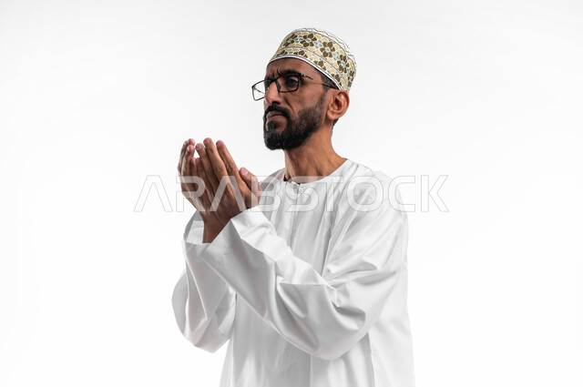 رفع اليدين للأعلى بهدف الدعاء والتضرع إلى الله تعالى،ايماءات التوسل والاسترحام، طلب المساعدة من المولى عز وجل، بورتريه لرجل عماني شايب يرتدي الدشداشة والكمه يدعي ويطلب المغفرة، خلفية بيضاء