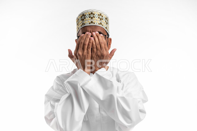 مسح الوجه باليدين دليل على الانتهاء من الدعاء، حركة اليد تدل على قول آمين، بورتريه لرجل عماني شايب يرتدي الدشداشة والكمه يدعي ويتوسل إلى الله، خلفية بيضاء