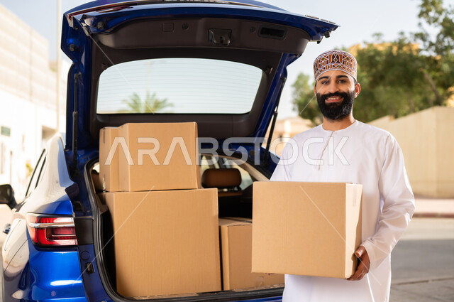 التسوق و شراء المنتجات عبر الانترنت, النظر إلى الكاميرا بإيماءات السعادة و السرور, خدمات الشحن السريعة و الآمنة, شاب عربي خليجي عماني يرتدي الدشداشة و الكمة يقف أمام سيارة التوصيل و يحمل بيده صندوق المشتريات