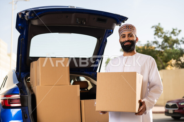 النظر إلى الكاميرا بإيماءات السعادة و السرور, التسوق و شراء المنتجات عبر الانترنت, خدمات الشحن السريعة و الآمنة, شاب عربي خليجي عماني يرتدي الدشداشة و الكمة يقف أمام سيارة التوصيل و يحمل بيده صندوق المشتريات
