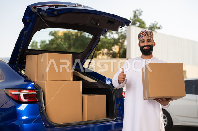 التسوق و شراء المنتجات عبر الانترنت, خدمات الشحن السريعة و الآمنة, النظر إلى الكاميرا بإيماءات السعادة و السرور, شاب عربي خليجي عماني يرتدي الدشداشة و الكمة يحمل بيده صندوق المشتريات و يرفع إصبع الإبهام بتعابير الجودة و الامتياز