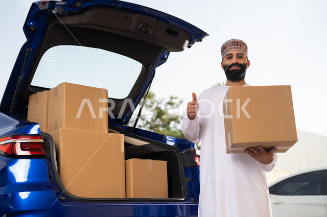 Shopping and purchasing products online, fast and safe shipping services, looking at the camera with gestures of happiness and pleasure, a young Arab Gulf Omani man wearing a dishdasha and a sleeve holding a shopping box in his hand and raising his thumb with expressions of quality and excellence.