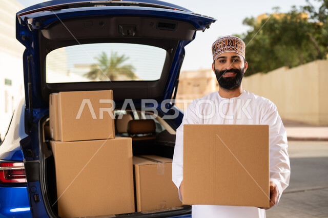 خدمات الشحن السريعة و الآمنة, النظر إلى الكاميرا بإيماءات السعادة و السرور, التسوق و شراء المنتجات عبر الانترنت, شاب عربي خليجي عماني يرتدي الدشداشة و الكمة يقف أمام سيارة التوصيل و يحمل بيده صندوق المشتريات