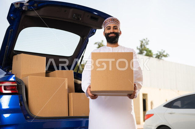 النظر إلى الكاميرا بإيماءات السعادة و السرور, خدمات الشحن السريعة و الآمنة, التسوق و شراء المنتجات عبر الانترنت, شاب عربي خليجي عماني يرتدي الدشداشة و الكمة يقف أمام سيارة التوصيل و يحمل بيده صندوق المشتريات