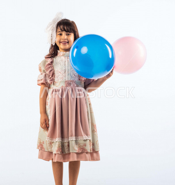 خلفية بيضاء لفتاه صغيرة سعودية خليجية مبتسمة وجميلة ، تنظر الى الكاميرا وتحمل بيدها  بالونين بلون أزرق وزهري بإيماءات وجه ويدين مختلفة تدل على السعادة , بورتريه لطفلة سعودية خليجية سعيدة ترتدي لباس العيد الانيق , الاحتفال بعيد الفطر السعيد ، عيد مبارك