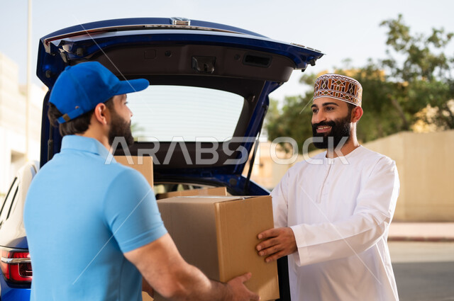 عمليات الشراء عبر الانترنت, طلب المنتجات أونلاين, خدمات شحن الطلبات للزبائن, مفهوم التسوق الالكتروني, شاب عربي خليجي عماني يرتدي الدشداشة و الكمة يقوم باستلام طرد المشتريات من مندوب التوصيل