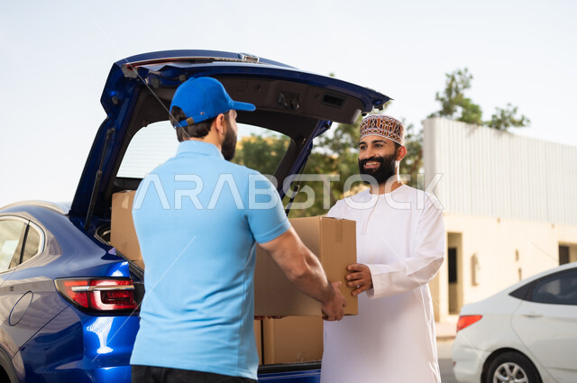 خدمات شحن الطلبات للزبائن, عمليات الشراء عبر الانترنت, طلب المنتجات أونلاين, مفهوم التسوق الالكتروني, شاب عربي خليجي عماني يرتدي الدشداشة و الكمة يقوم باستلام طرد المشتريات من مندوب التوصيل