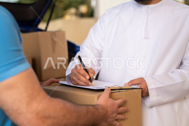 طلب المنتجات عبر الانترنت, مفهوم التسوق الالكتروني, خدمات الشحن ذات الجودة العالية, عمليات الشراء أونلاين, صورة مقربة ليد شاب عربي خليجي عماني يرتدي الدشداشة و الكمة يقوم بالتوقيع على أوراق و مستندات للتأكيد على استلام الطرد، مندوب يقوم بتوصيل طلبات الزبائن