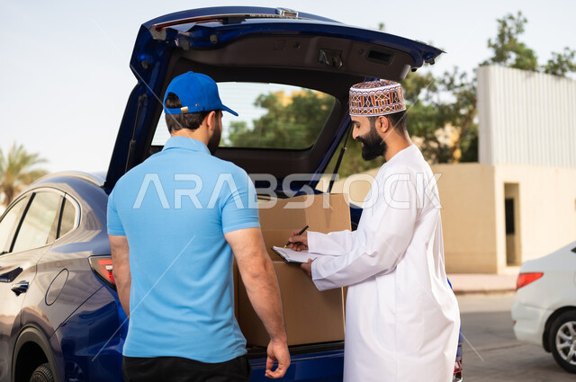 مفهوم التسوق الالكتروني, خدمات الشحن ذات الجودة العالية, عملية شراء المنتجات أونلاين, شاب عربي خليجي عماني يرتدي الدشداشة و الكمة يقوم بالتوقيع على أوراق و مستندات للتأكيد على استلام الطرد، مندوب يقوم بتوصيل طلبات الزبائن