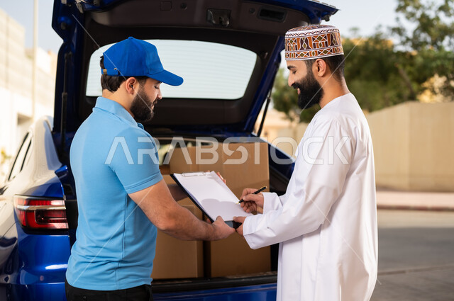 عملية شراء المنتجات أونلاين, خدمات الشحن ذات الجودة العالية, مفهوم التسوق الالكتروني, شاب عربي خليجي عماني يرتدي الدشداشة و الكمة يقوم بالتوقيع على أوراق و مستندات للتأكيد على استلام الطرد، مندوب يقوم بتوصيل طلبات الزبائن