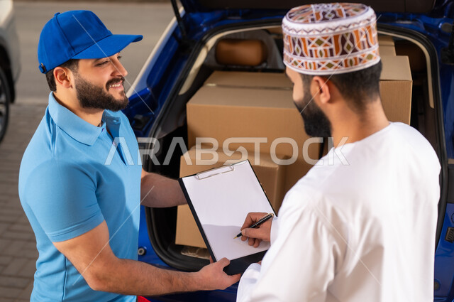 عملية شراء المنتجات أونلاين, خدمات الشحن ذات الجودة العالية, مفهوم التسوق الالكتروني, شاب عربي خليجي عماني يرتدي الدشداشة و الكمة يقوم بالتوقيع على أوراق و مستندات للتأكيد على استلام الطرد، مندوب يقوم بتوصيل طلبات الزبائن