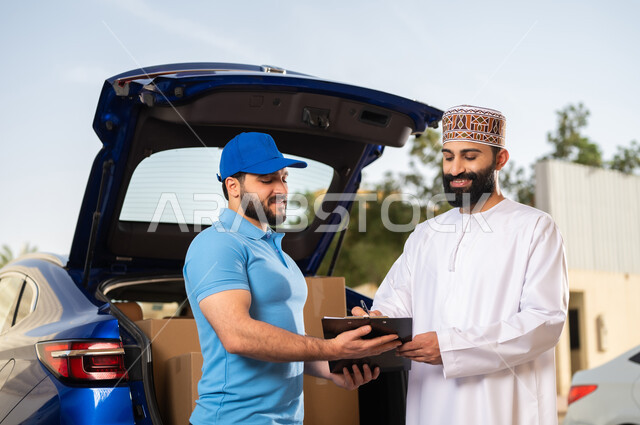 خدمات الشحن ذات الجودة العالية, عمليات الشراء أونلاين, مفهوم التسوق الالكتروني, شاب عربي خليجي عماني يرتدي الدشداشة و الكمة يقوم بالتوقيع على أوراق و مستندات للتأكيد على استلام الطرد، مندوب يقوم بتوصيل طلبات الزبائن