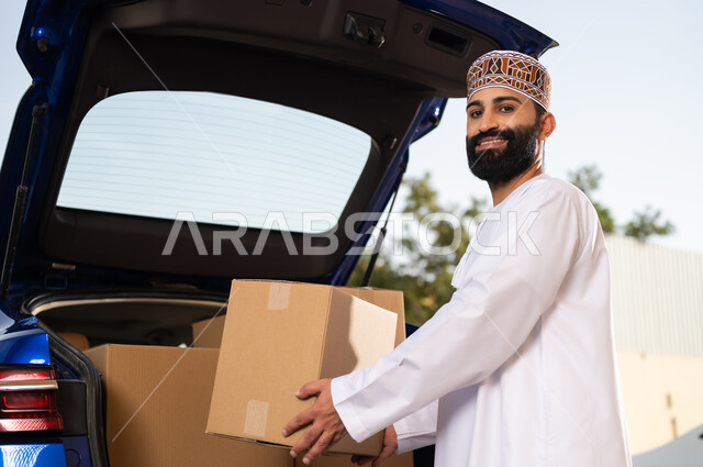 خدمات التوصيل السريع, شراء المنتجات أونلاين, النظر إلى الكاميرا بإيماءات السعادة و السرور, شاب عربي خليجي عماني يرتدي الدشداشة و الكمة يقوم بشحن الطرود بالسيارة, مفهوم التسوق الالكتروني