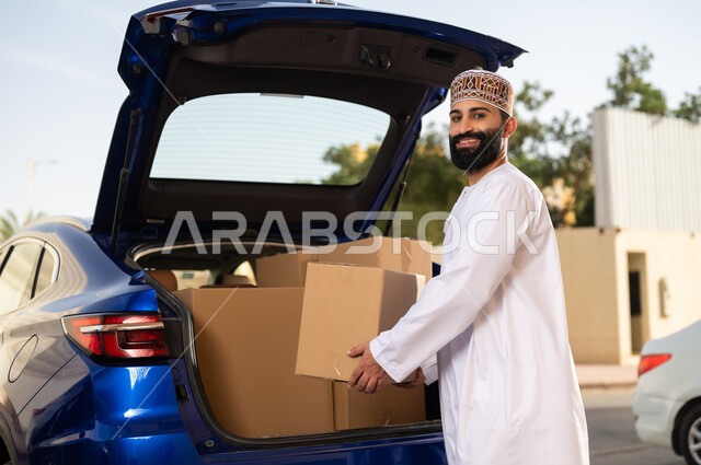 خدمات التوصيل السريع, شراء المنتجات أونلاين, النظر إلى الكاميرا بإيماءات السعادة و السرور, شاب عربي خليجي عماني يرتدي الدشداشة و الكمة يقوم بشحن الطرود بالسيارة, مفهوم التسوق الالكتروني