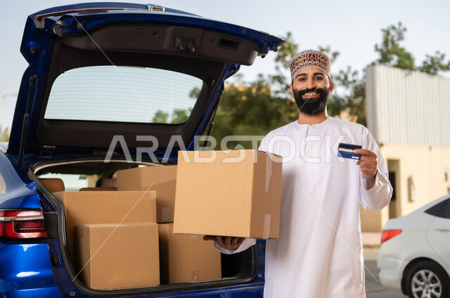 عمليات الدفع عن بعد, شراء المنتجات أونلاين, مفهوم التسوق الالكتروني, شاب عربي خليجي عماني يرتدي الدشداشة و الكمة يحمل بيده صندوق و بطاقة الائتمان البنكية, خدمات التوصيل السريع