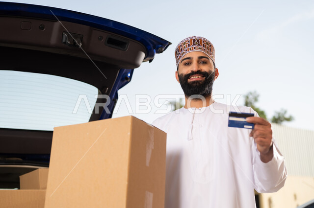 عمليات الدفع عن بعد, شراء المنتجات أونلاين, مفهوم التسوق الالكتروني, شاب عربي خليجي عماني يرتدي الدشداشة و الكمة يحمل بيده صندوق و بطاقة الائتمان البنكية, خدمات التوصيل السريع