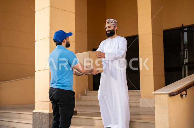 Ordering products online, the concept of electronic shopping and online orders, an electronic purchase process, express delivery services, an Arab Gulf Omani young man wearing a dishdasha and a sleeve receiving a package, a representative delivering a shopping box
