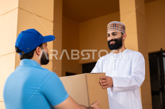 طلب المنتجات أونلاين, مفهوم التسوق الالكتروني و الطلبات أونلاين, عملية شراء الكترونية، خدمات التوصيل السريع, شاب عربي خليجي عماني يرتدي الدشداشة و الكمة يقوم باستلام طرد, مندوب يقوم بتسليم صندوق مشتريات