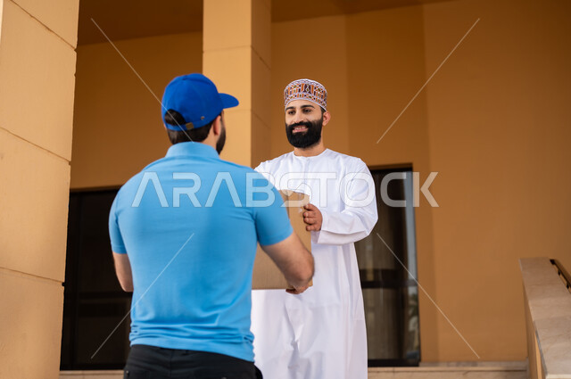 مفهوم التسوق الالكتروني و الطلبات أونلاين, عملية شراء الكترونية، خدمات التوصيل السريع, شاب عربي خليجي عماني يرتدي الدشداشة و الكمة يقوم باستلام طرد, مندوب يقوم بتسليم صندوق مشتريات