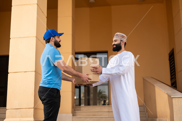 مفهوم التسوق الالكتروني و الطلبات أونلاين, عملية شراء الكترونية، خدمات التوصيل السريع, شاب عربي خليجي عماني يرتدي الدشداشة و الكمة يقوم باستلام طرد, مندوب يقوم بتسليم صندوق مشتريات