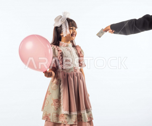 خلفية بيضاء لفتاه صغيرة سعودية خليجية مبتسمة وجميلة ، تحمل بيدها بالون زهري وتنظر للعيدية بإيماءات وجه ويدين مختلفة تدل على السعادة , بورتريه لطفلة سعودية خليجية سعيدة ترتدي لباس العيد الانيق, الاحتفال بعيد الفطر السعيد , تقديم العيدية للأطفال