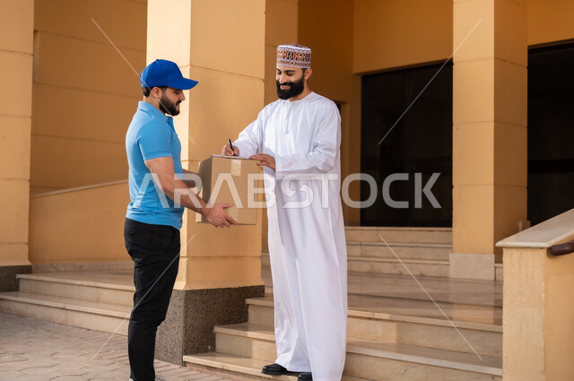 خدمات الشحن ذات الجودة العالية, مفهوم التسوق الالكتروني, شاب عربي خليجي عماني يرتدي الدشداشة و الكمة يقوم بالتوقيع على أوراق و مستندات للتأكيد على استلام الطرد، عملية توصيل الطلبات للزبائن