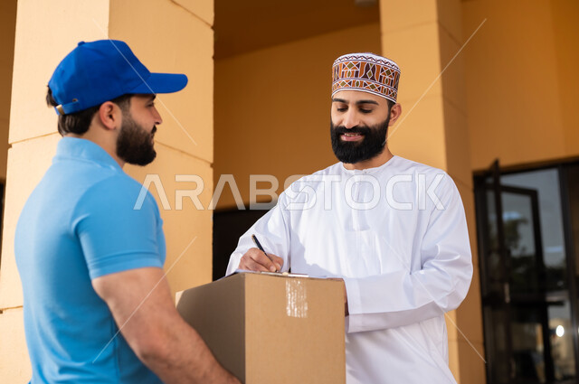 خدمات الشحن ذات الجودة العالية, مفهوم التسوق الالكتروني, شاب عربي خليجي عماني يرتدي الدشداشة و الكمة يقوم بالتوقيع على أوراق و مستندات للتأكيد على استلام الطرد، عملية توصيل الطلبات للزبائن